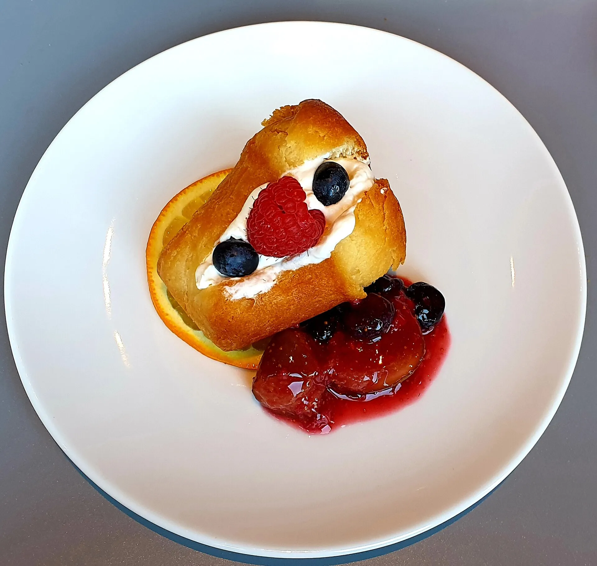 Baba bouchon garni de crème, de myrtilles, d'une framboise et d'une tranche d'orange, vu de dessus