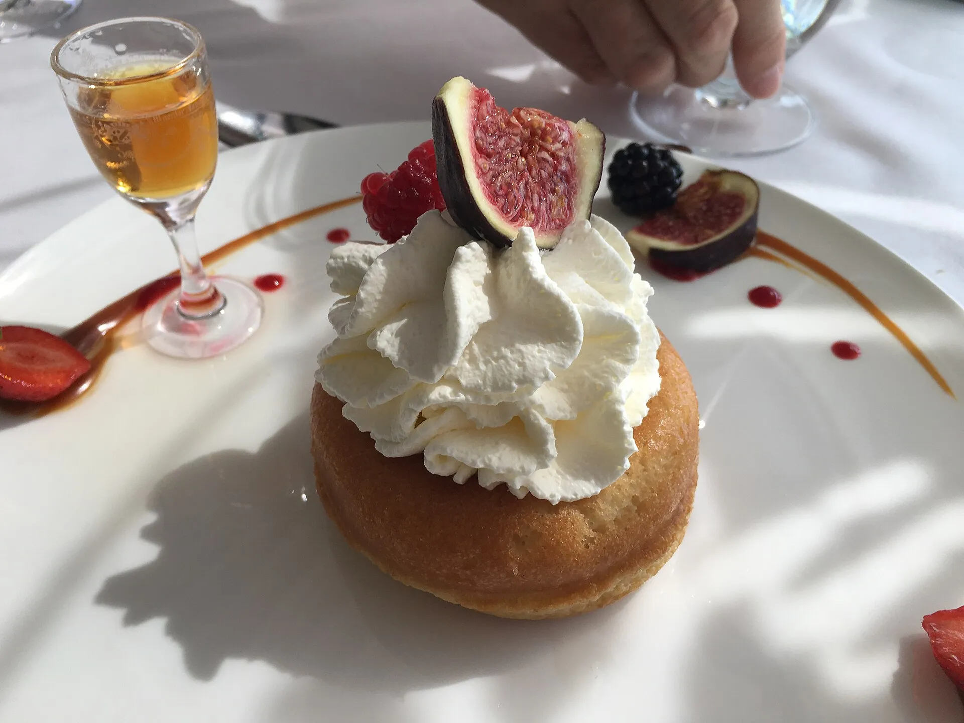 Baba à la chantilly garni de figues et de mûres, un petit verre de rhum ambré posé à côté sur la nappe blanche